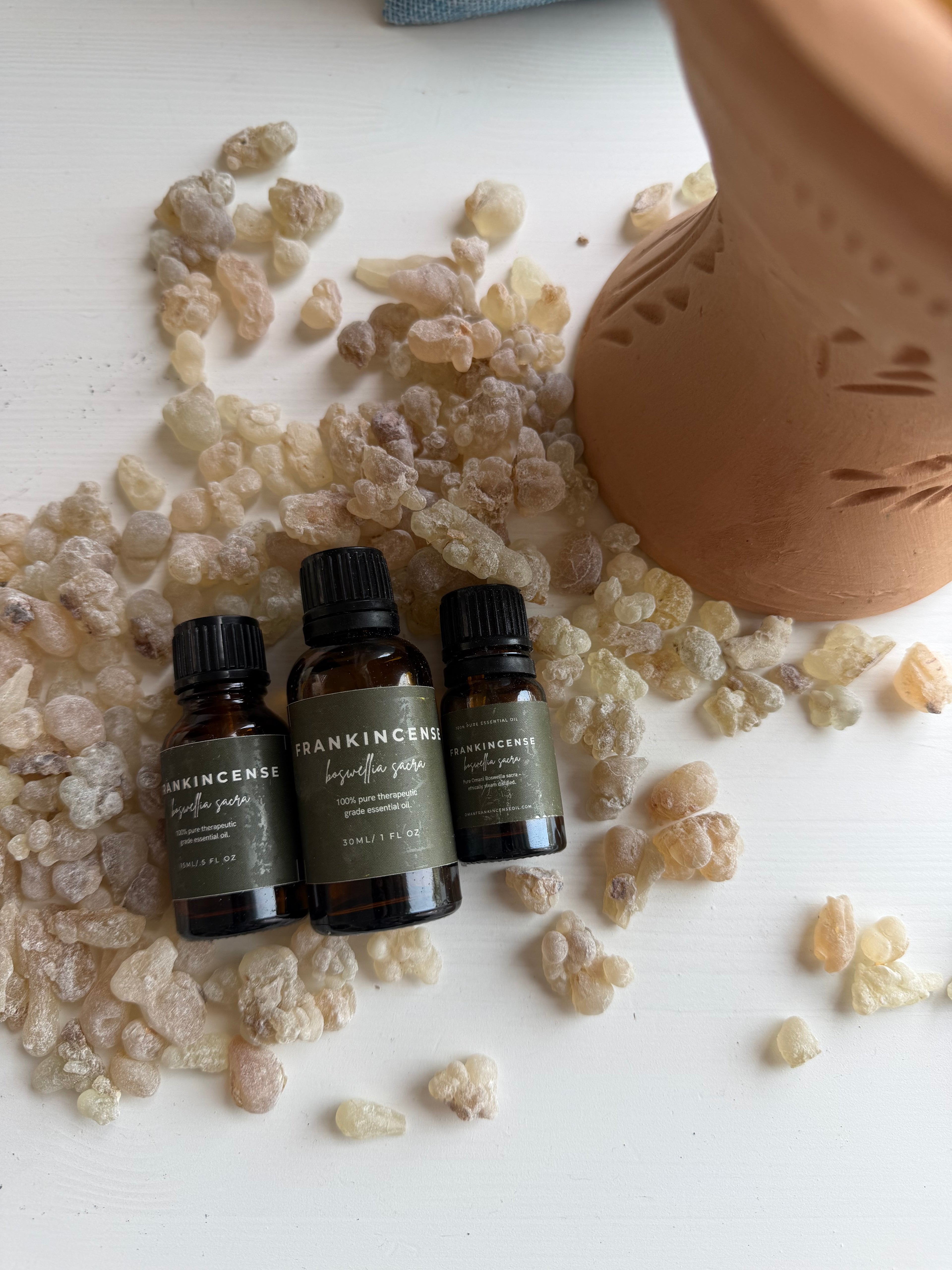 Three small amber glass essential oil bottles with black caps and dark olive labels are arranged on a white surface surrounded by resin pieces and a terracotta pot. The labels show the brand Frankincense and scent names including Immense Power.