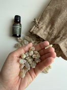 A hand holding Hojari Frankincense resin spilled from a brown burlap bag, with a small dark amber bottle labeled 'Frankincense' on a white surface.