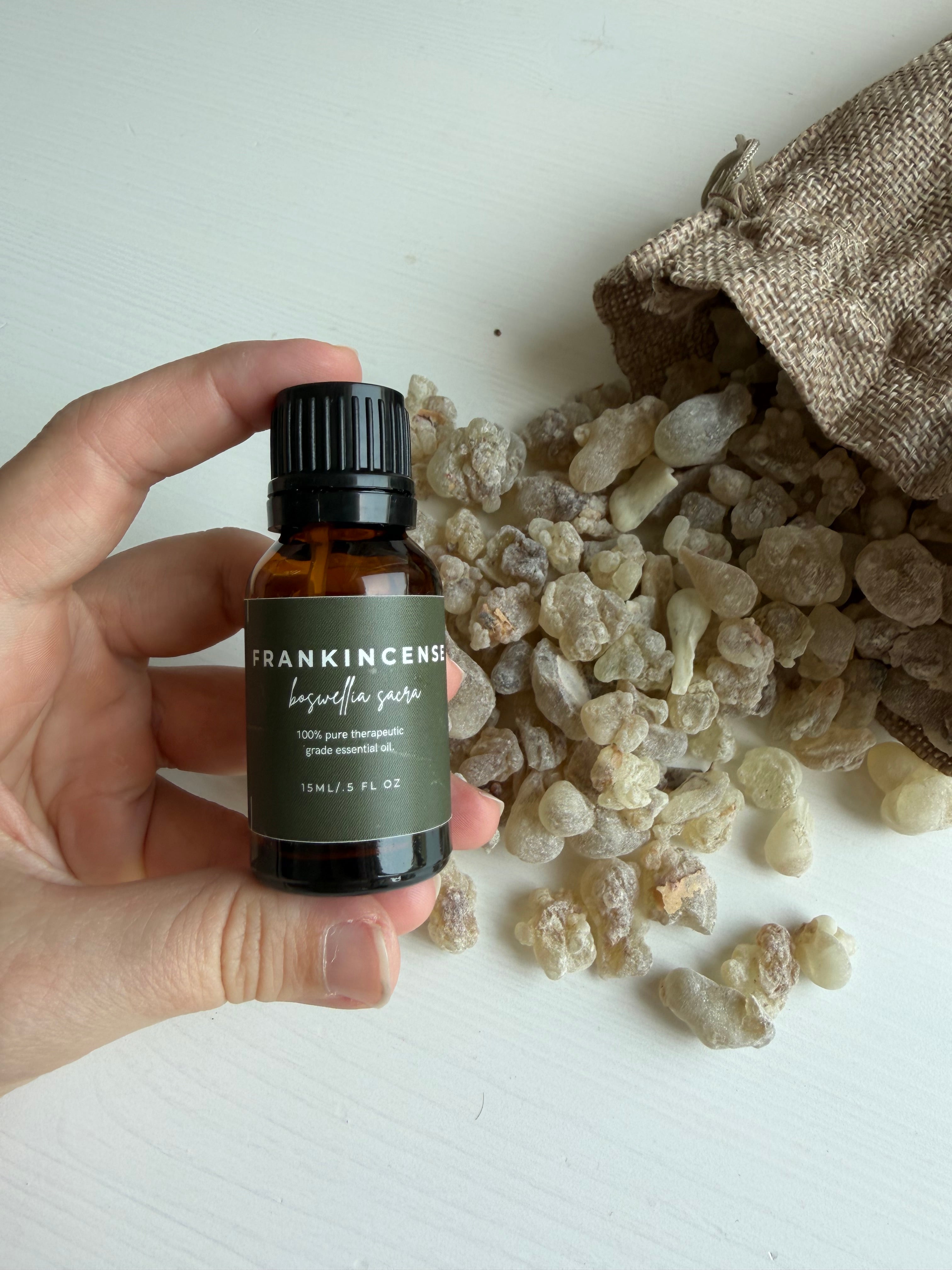 A hand holds a small amber glass bottle labeled Frankincense with a dark olive green label and black cap, set against a white background with raw unpolished resin pieces and a burlap sack.