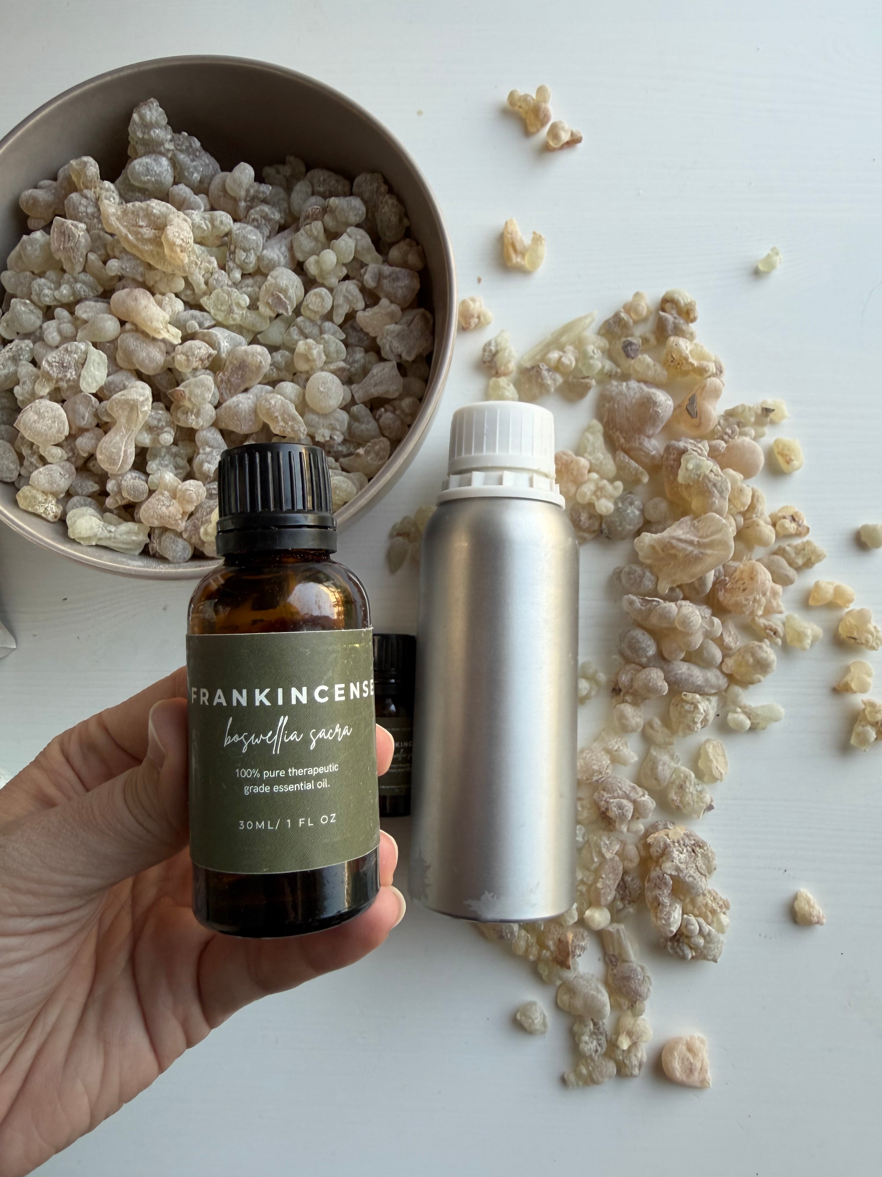 A hand holds a small amber bottle labeled Frankincense essential oil oil next to a silver oil canister and a bowl of natural resin pieces on a white surface.