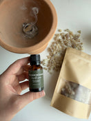 A hand holds a small amber bottle labeled Frankincense over a terracotta bowl with smoke rising, with a kraft pouch and scattered resin pieces on a white surface.