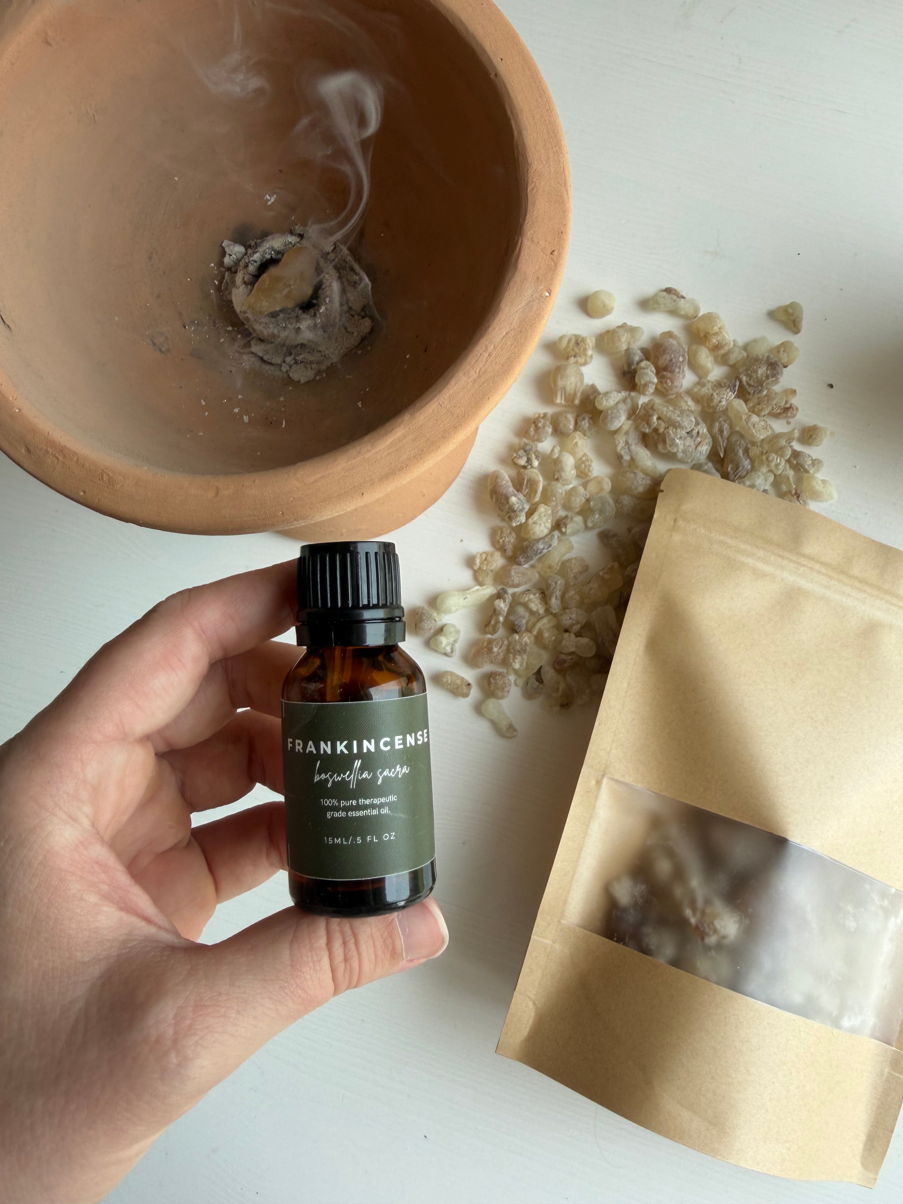 A hand holds a small amber bottle labeled Frankincense over a terracotta bowl with smoke rising, with a kraft pouch and scattered resin pieces on a white surface.